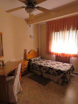 Un dormitorio con cama, escritorio, silla, ventilador de techo y ventana con cortinas.