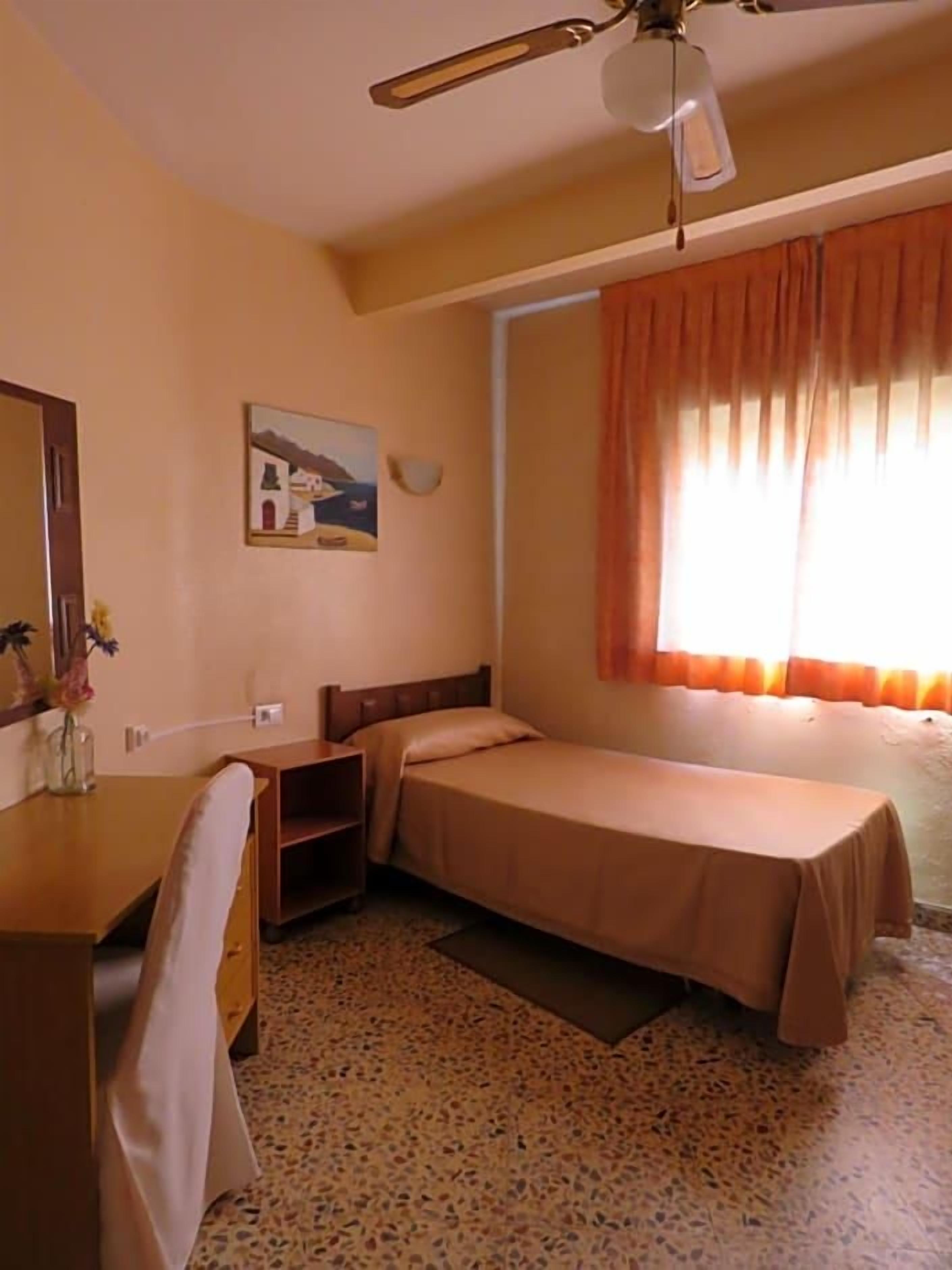 A small, single-bed room with a desk, chair, and a ceiling fan.