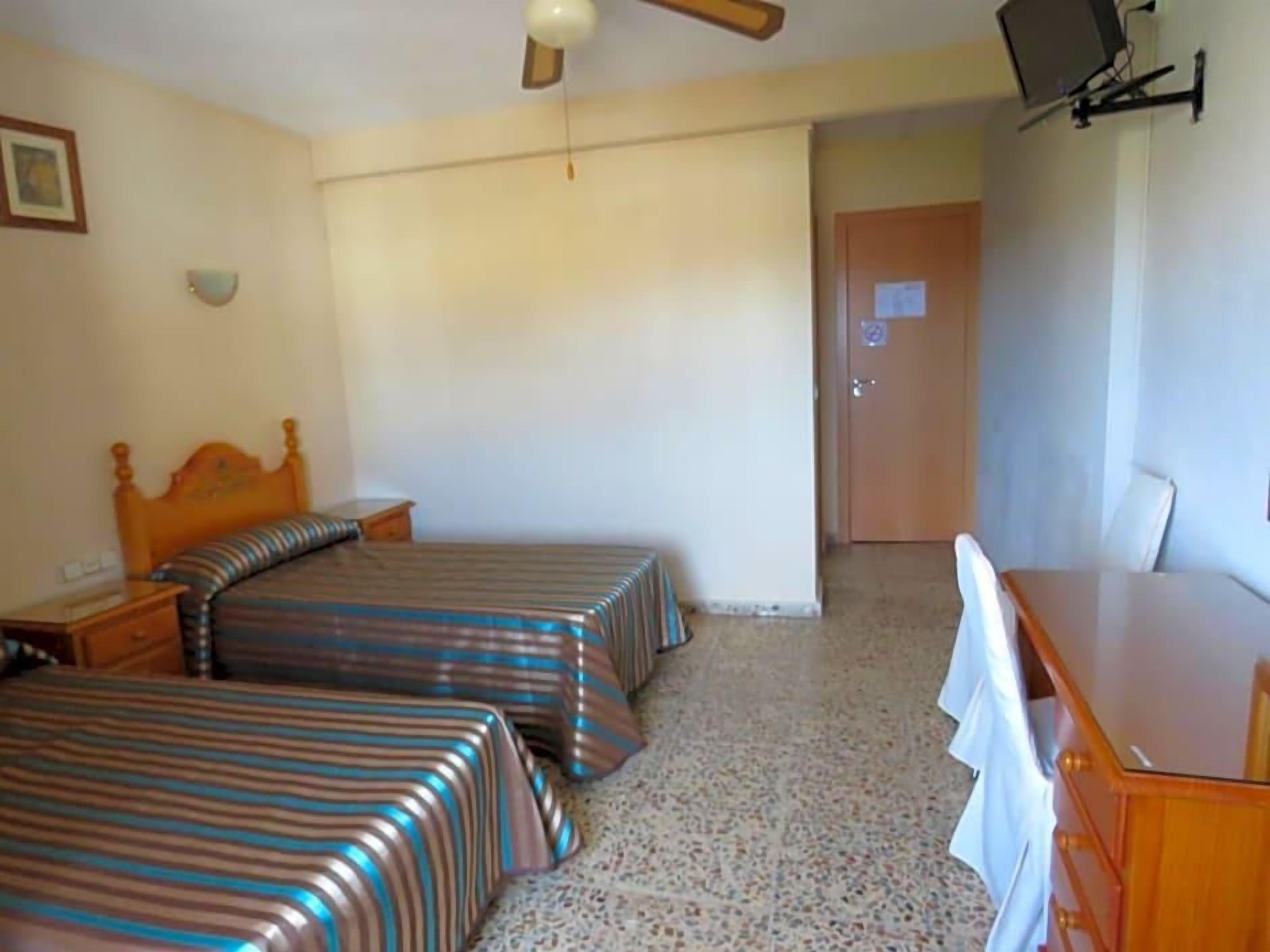 Un dormitorio con dos camas, un aparador de madera, un escritorio, un ventilador de techo y una puerta.