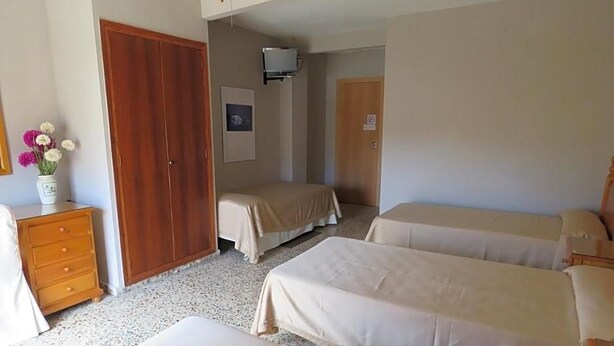 A hotel room with three beds, a wooden wardrobe, a desk with a vase of flowers, and a television on the wall.