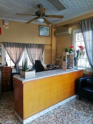 A reception area with a desk, a ceiling fan, a window with curtains, a potted plant, and a tiled floor. - Hotel Alain (Silla)