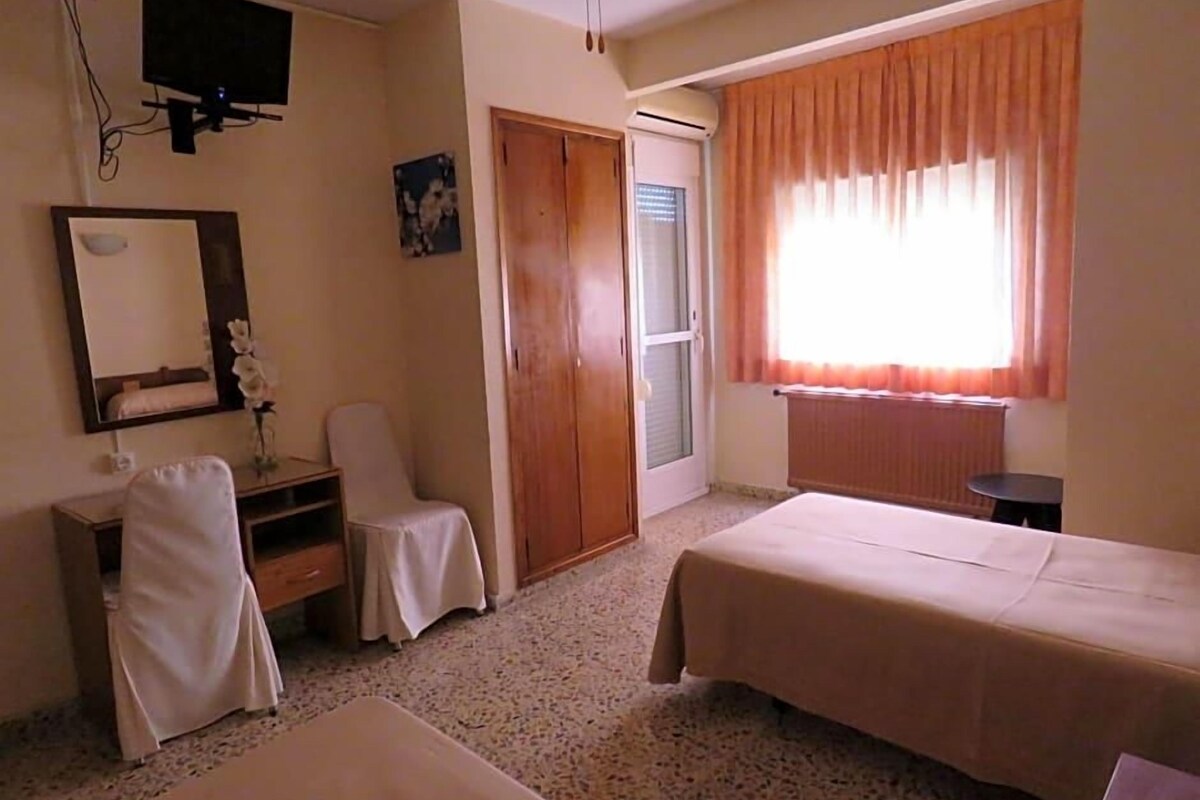 A room with two beds, a chair, a desk, a mirror, a television, and a window with curtains.