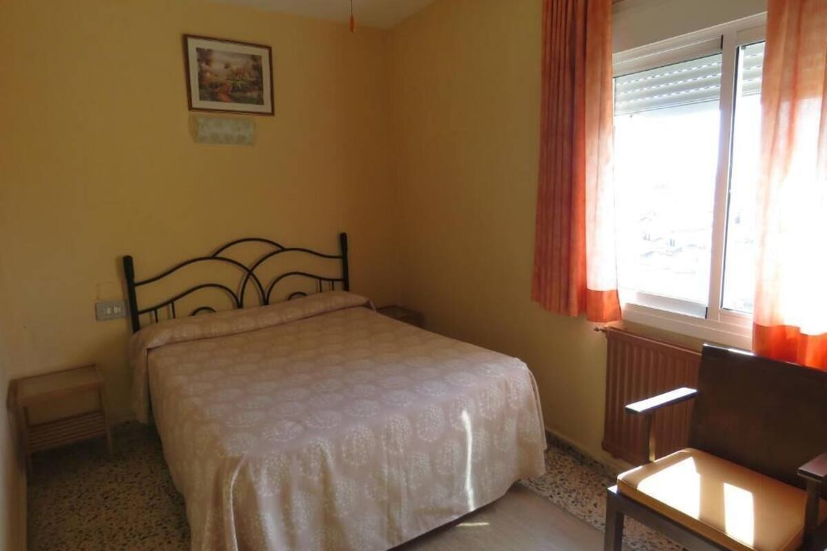 A bedroom with a bed, a chair, a radiator, a window with curtains, and a picture on the wall.