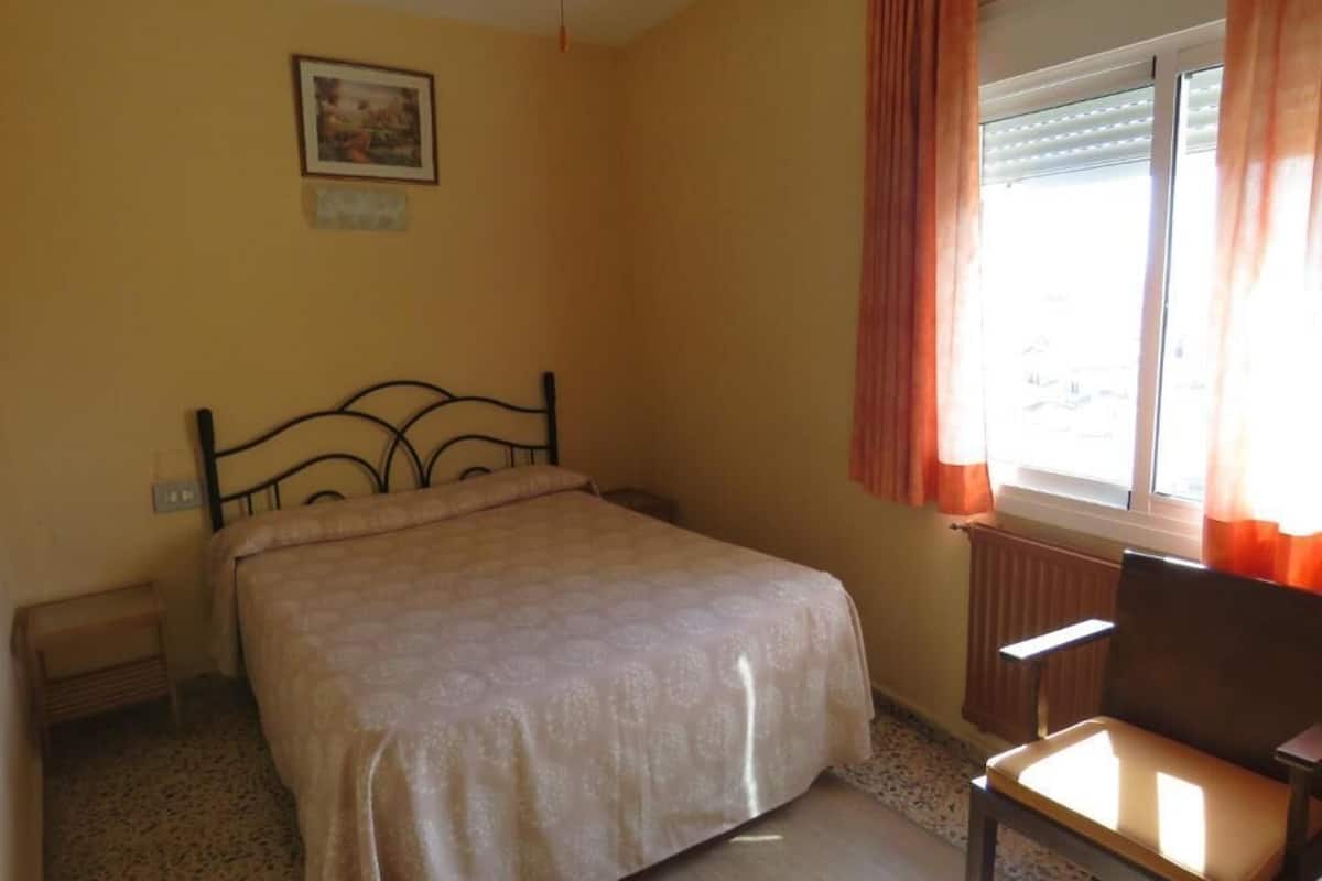 Une chambre à coucher avec un lit, une chaise, un radiateur, une fenêtre avec des rideaux et un tableau au mur.