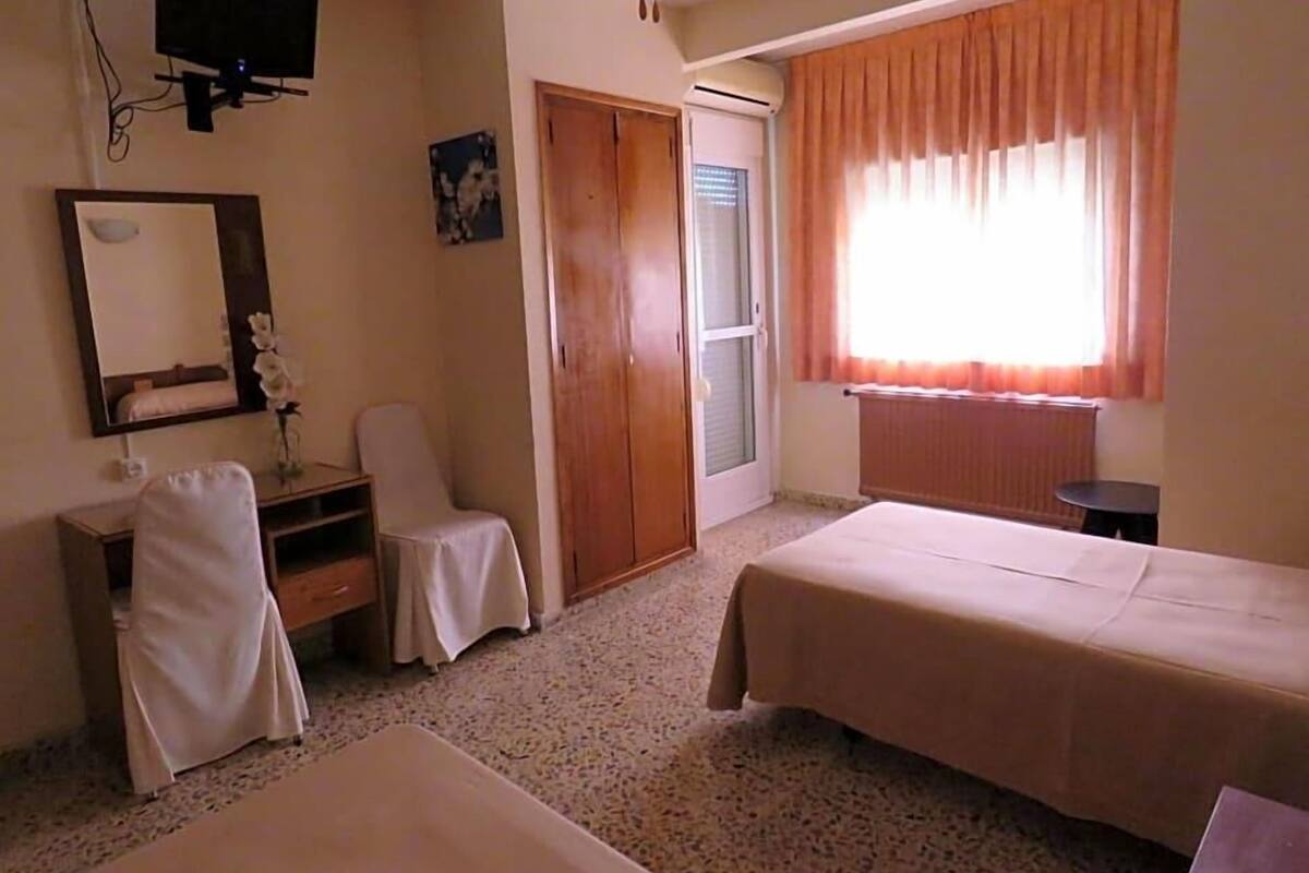 A room with two beds, a chair, a desk, a mirror, a television, and a window with curtains.