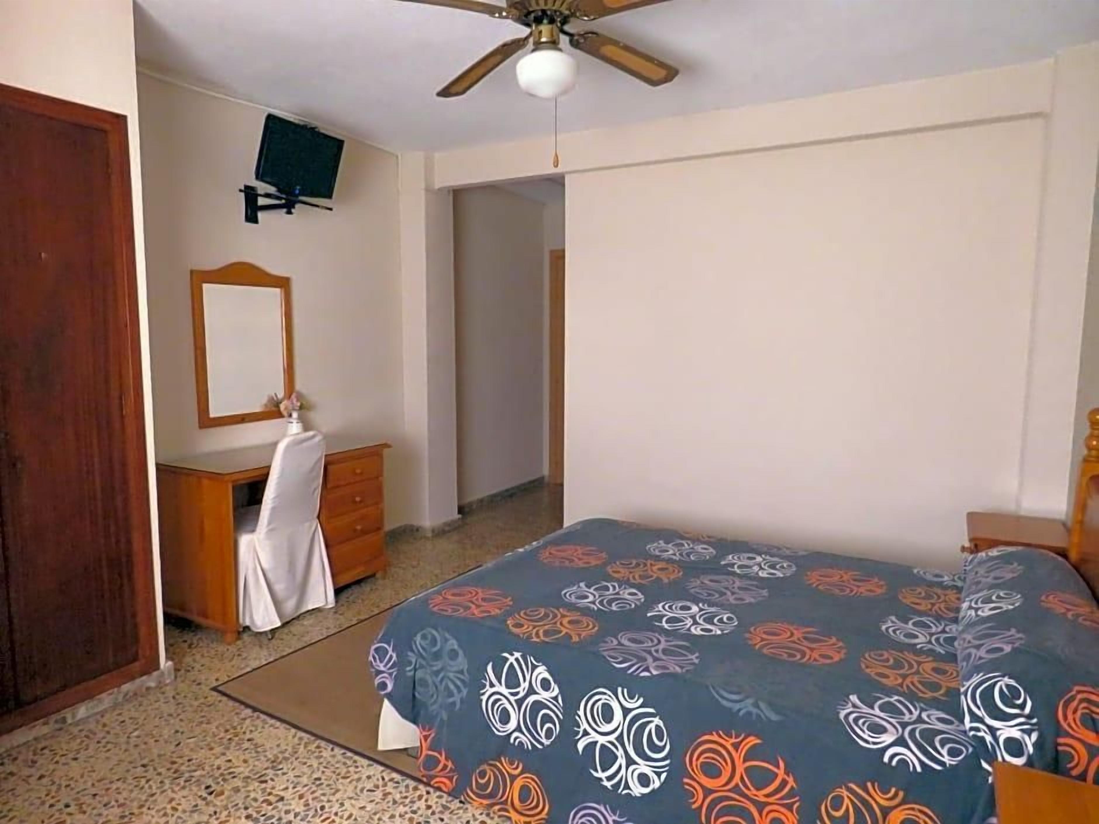 A bedroom with a bed, a dresser, a mirror, a ceiling fan, and a television on the wall.