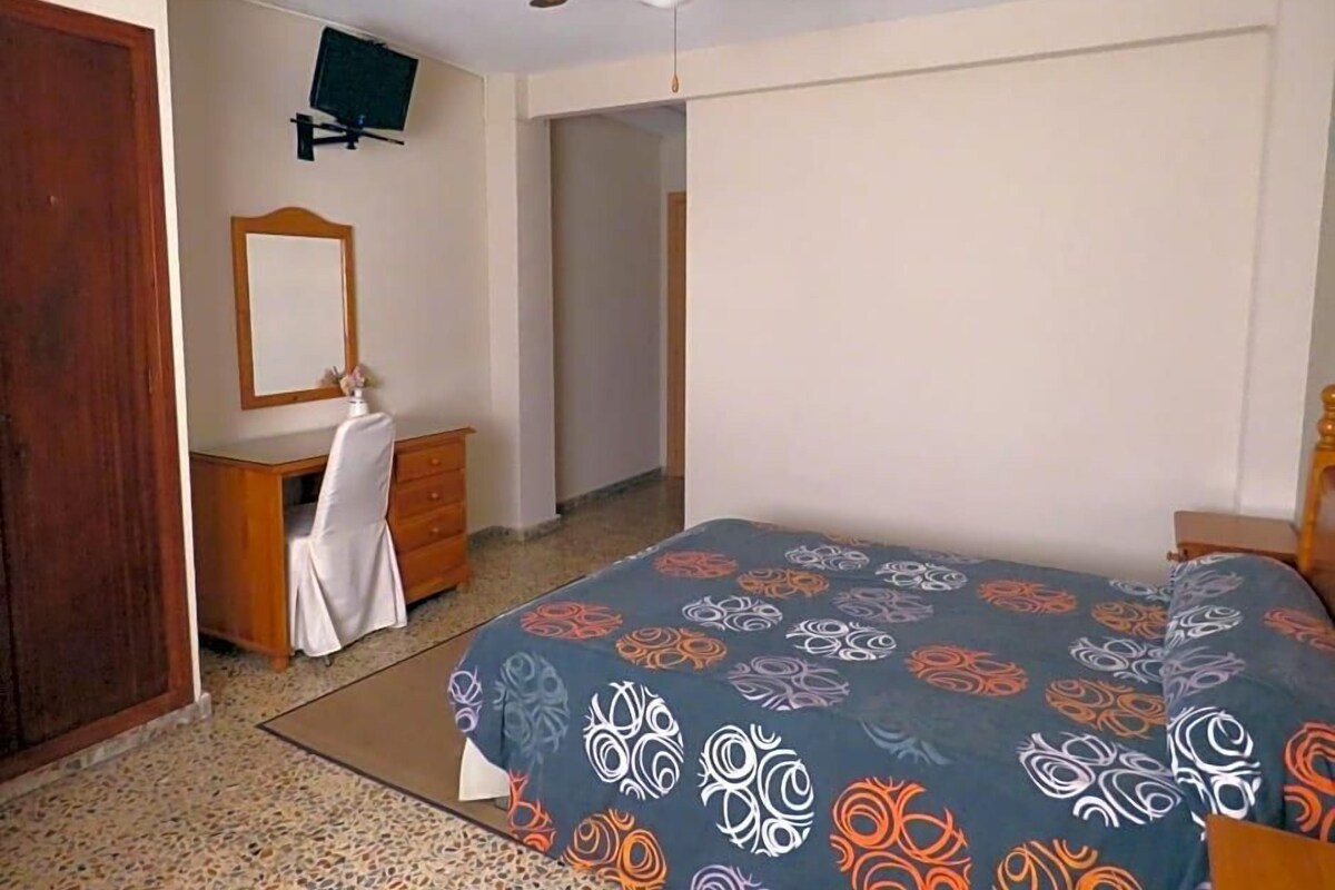 A bedroom with a bed, a dresser, a mirror, a ceiling fan, and a television on the wall.