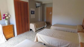 A hotel room with three beds, a wooden wardrobe, a desk with a vase of flowers, and a television on the wall.