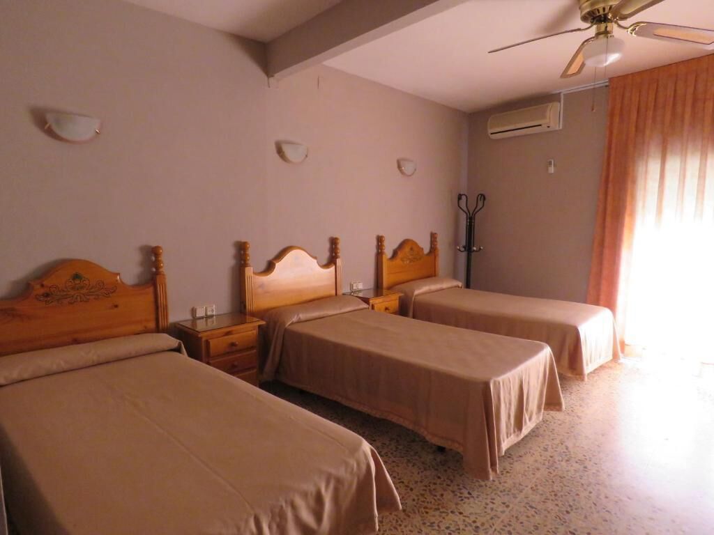 Une chambre d’hôtel avec trois lits, un ventilateur de plafond, un porte-manteau et une fenêtre avec des rideaux.