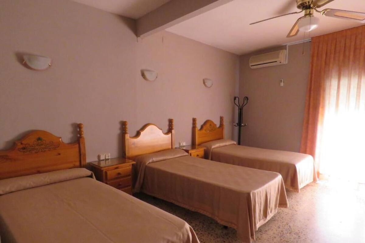 A hotel room with three beds, a ceiling fan, a coat rack, and a window with curtains.