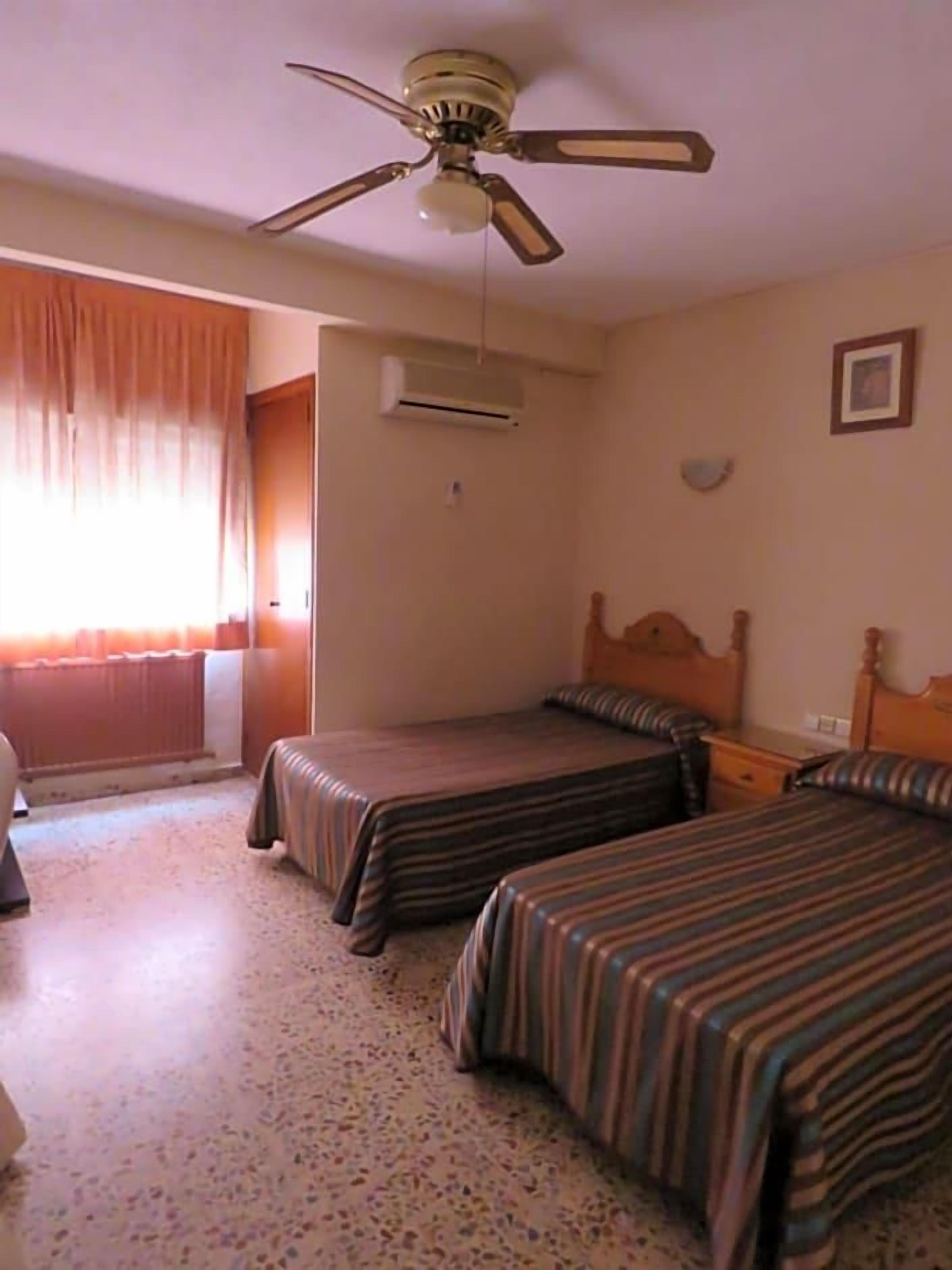 Una habitación de hotel con dos camas, un ventilador de techo, un equipo de aire acondicionado, una ventana con cortinas y una mesita de noche.
