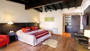 A modern hotel room with a large bed, wooden flooring, a red armchair, a desk with a lamp, and a view of the kitchen. - Monasterio Hotel Boutique (San Carlos de Bariloche)