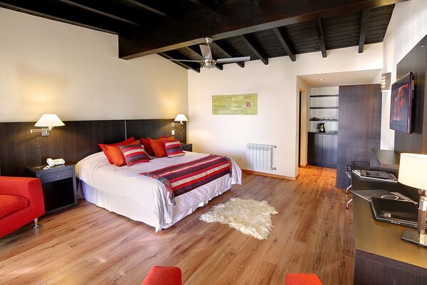 A modern hotel room with a large bed, wooden flooring, a red armchair, a desk with a lamp, and a view of the kitchen. - Monasterio Hotel Boutique (San Carlos de Bariloche)