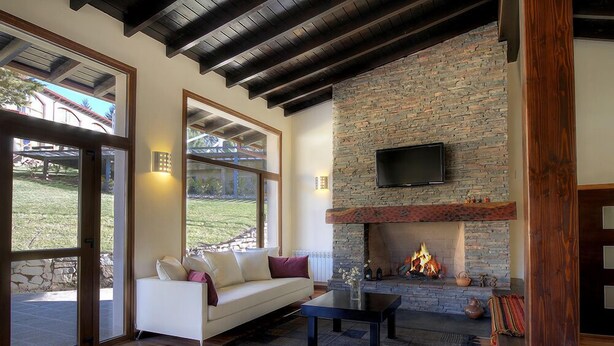 A modern living room with a stone fireplace, a flat-screen TV mounted above it, and a white sofa with cushions. - Monasterio Hotel Boutique (San Carlos de Bariloche)
