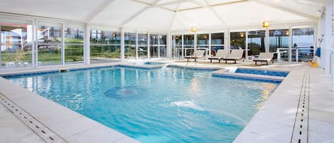 An indoor swimming pool with clear glass walls, lounge chairs, and a covered roof.