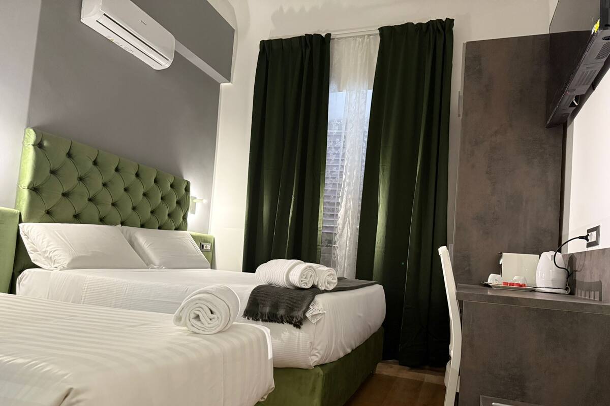 A hotel room with two beds, a green headboard, a desk with a kettle, and a window with curtains.
