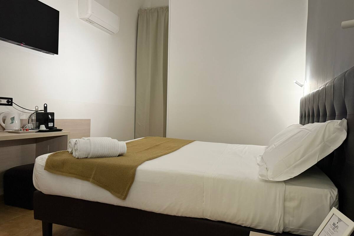 A neatly made bed with white linens and a brown throw, a bedside table with a coffee maker, and a wall-mounted air conditioner.