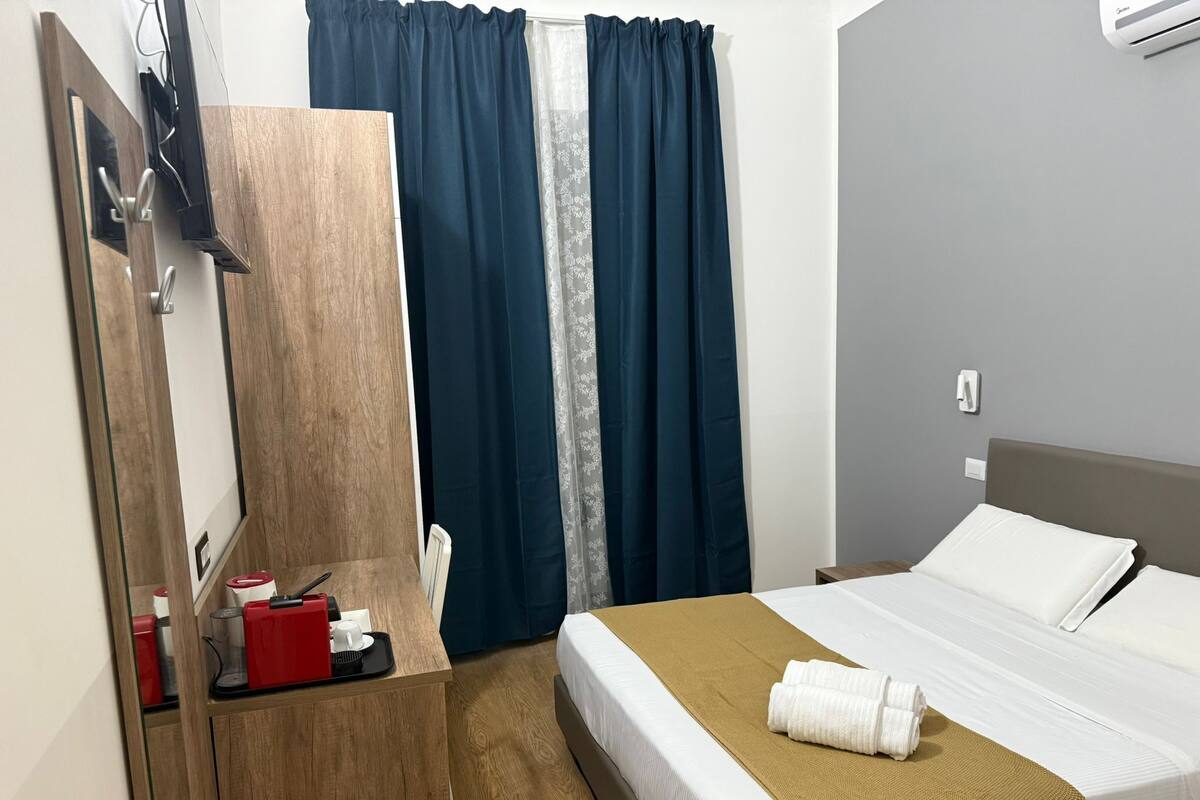 A hotel room with a bed, a wooden desk, a mirror, a TV, and a window with curtains.