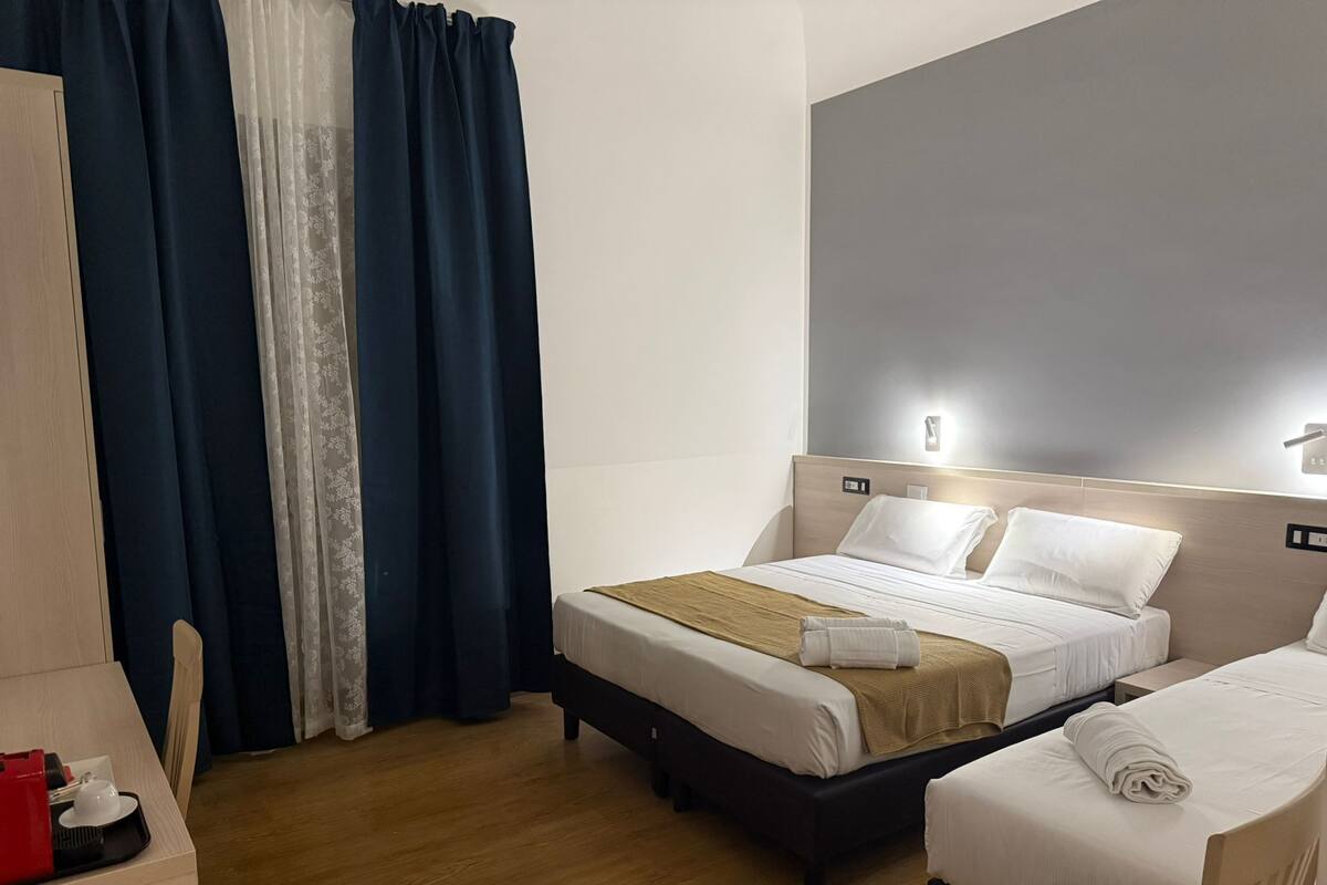 A hotel room with two beds, a wooden wardrobe, and a desk with a red object on it.