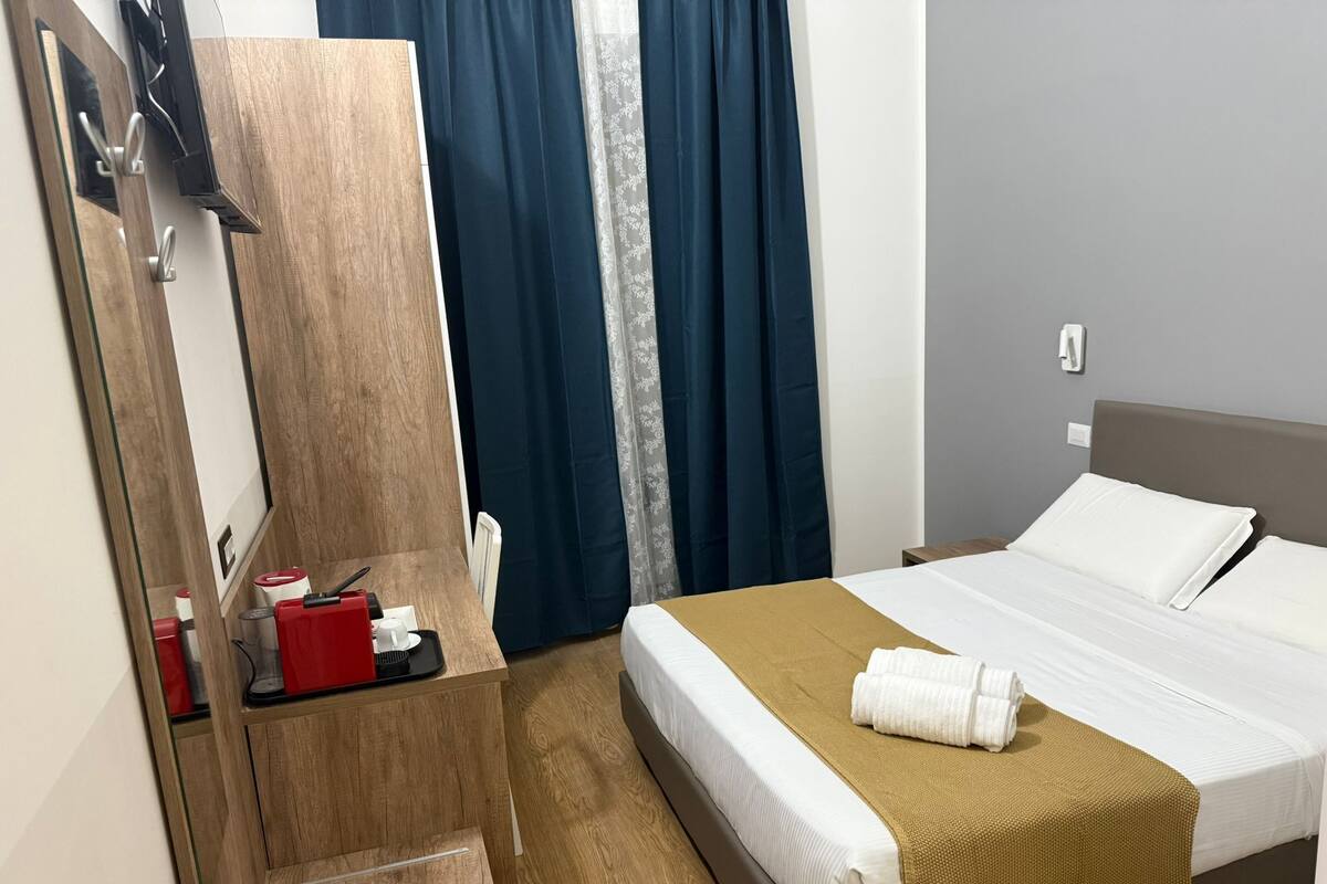 A hotel room with a bed, a wooden nightstand, a mirror, a small table with toiletries, and a window with curtains.