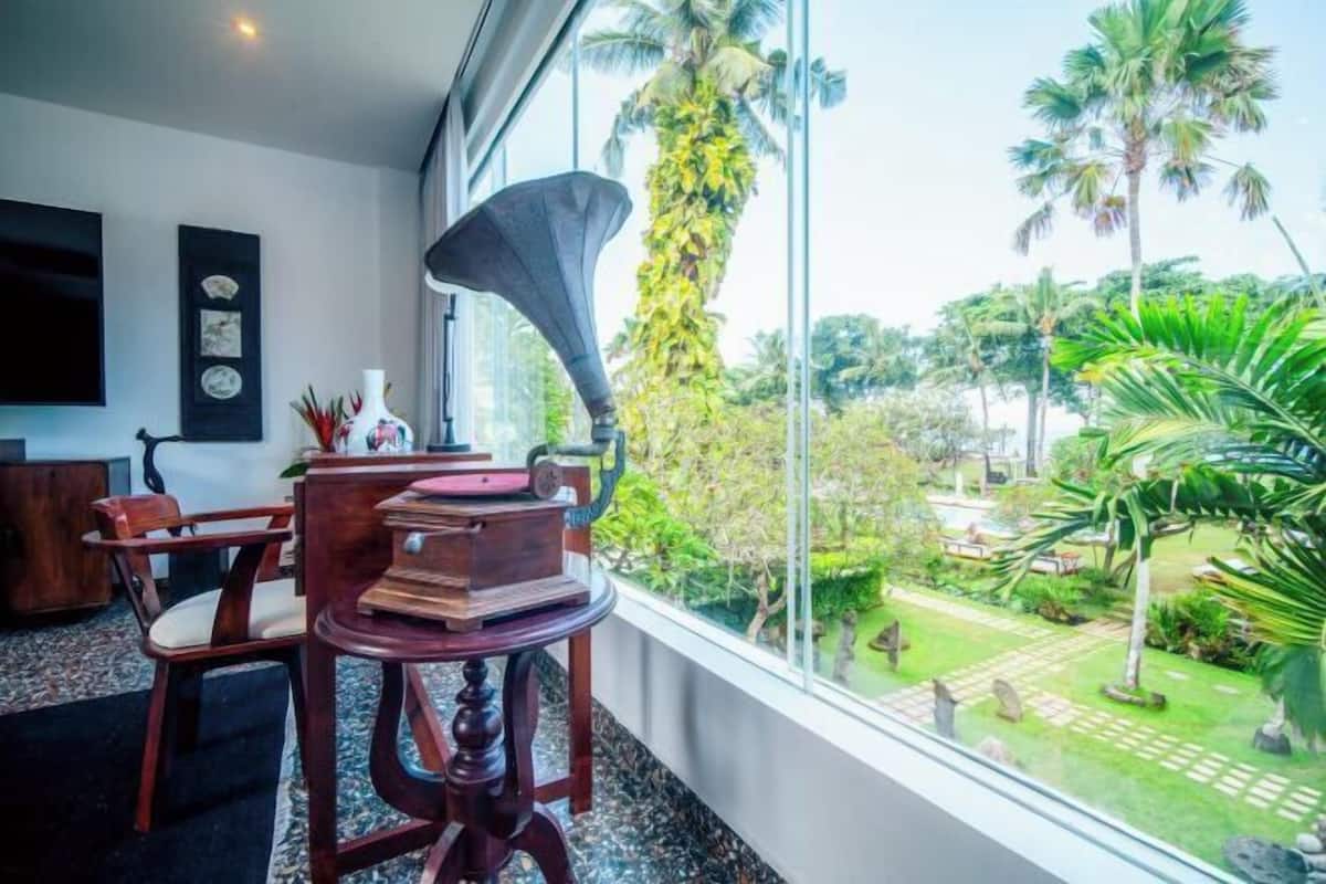 A room with a large window, a wooden table, a vintage gramophone, and a view of a tropical garden.
