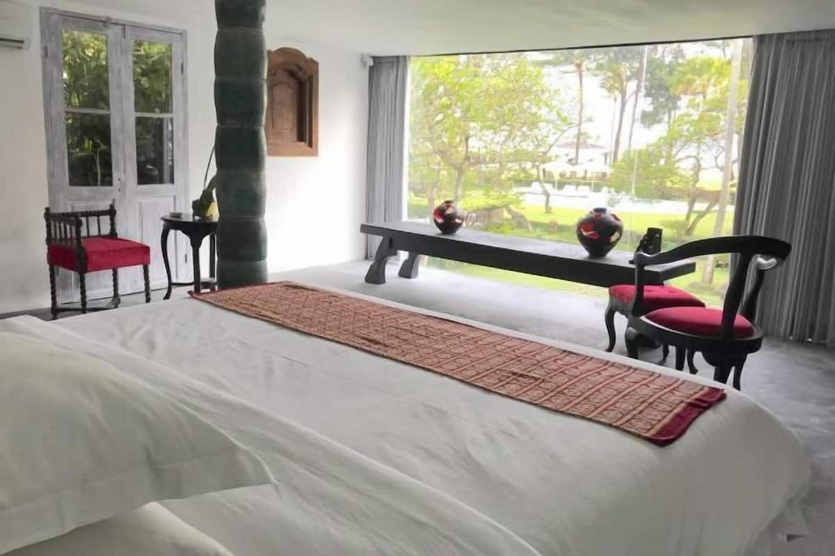 A bedroom with a bed, a window with a view of greenery, a bench, and two chairs.