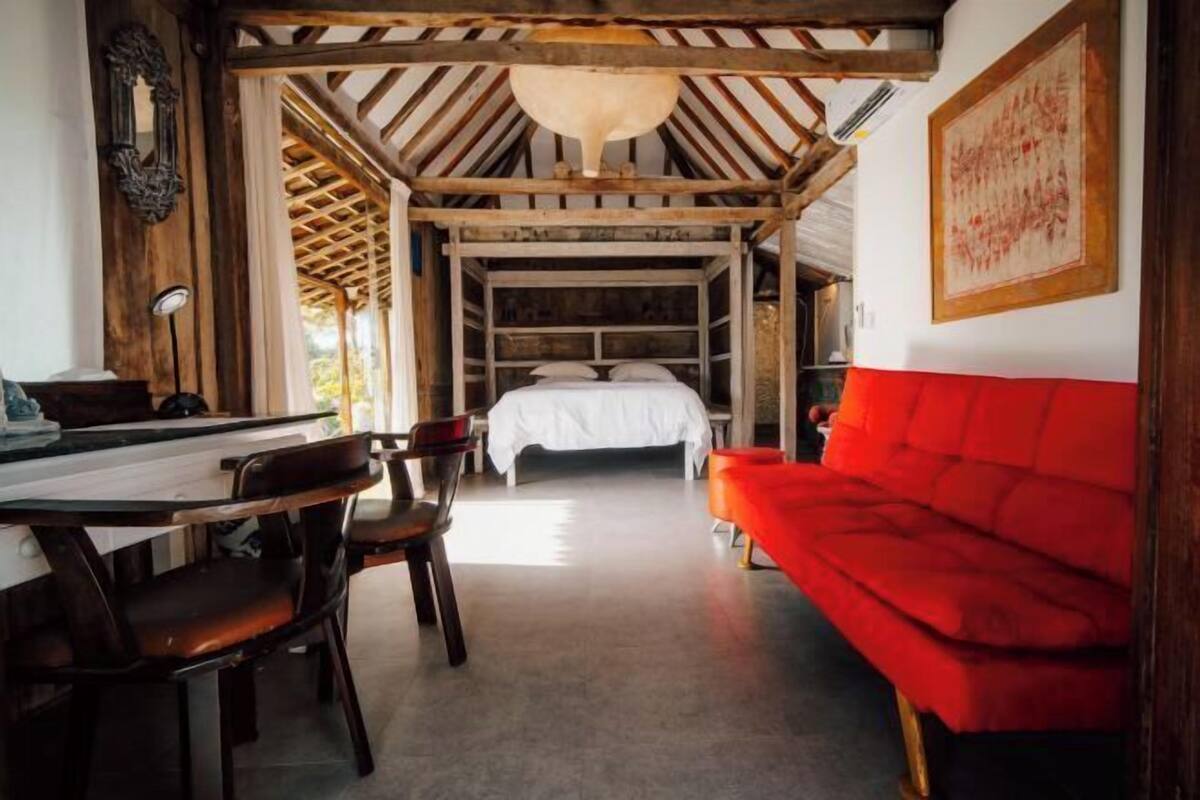 A room with a bed, a red sofa, a wooden table with chairs, and a ceiling with exposed beams.