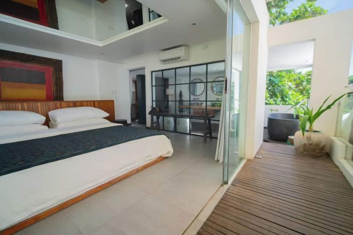 A modern bedroom with a large bed, a wooden floor, and a glass door leading to a balcony with a potted plant.