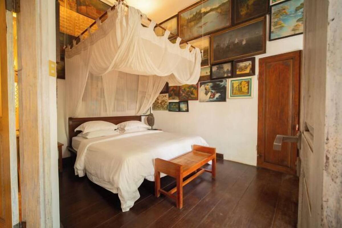 A bedroom with a bed, a wooden bench, a bedside table, a bedside lamp, and a wall with framed pictures.