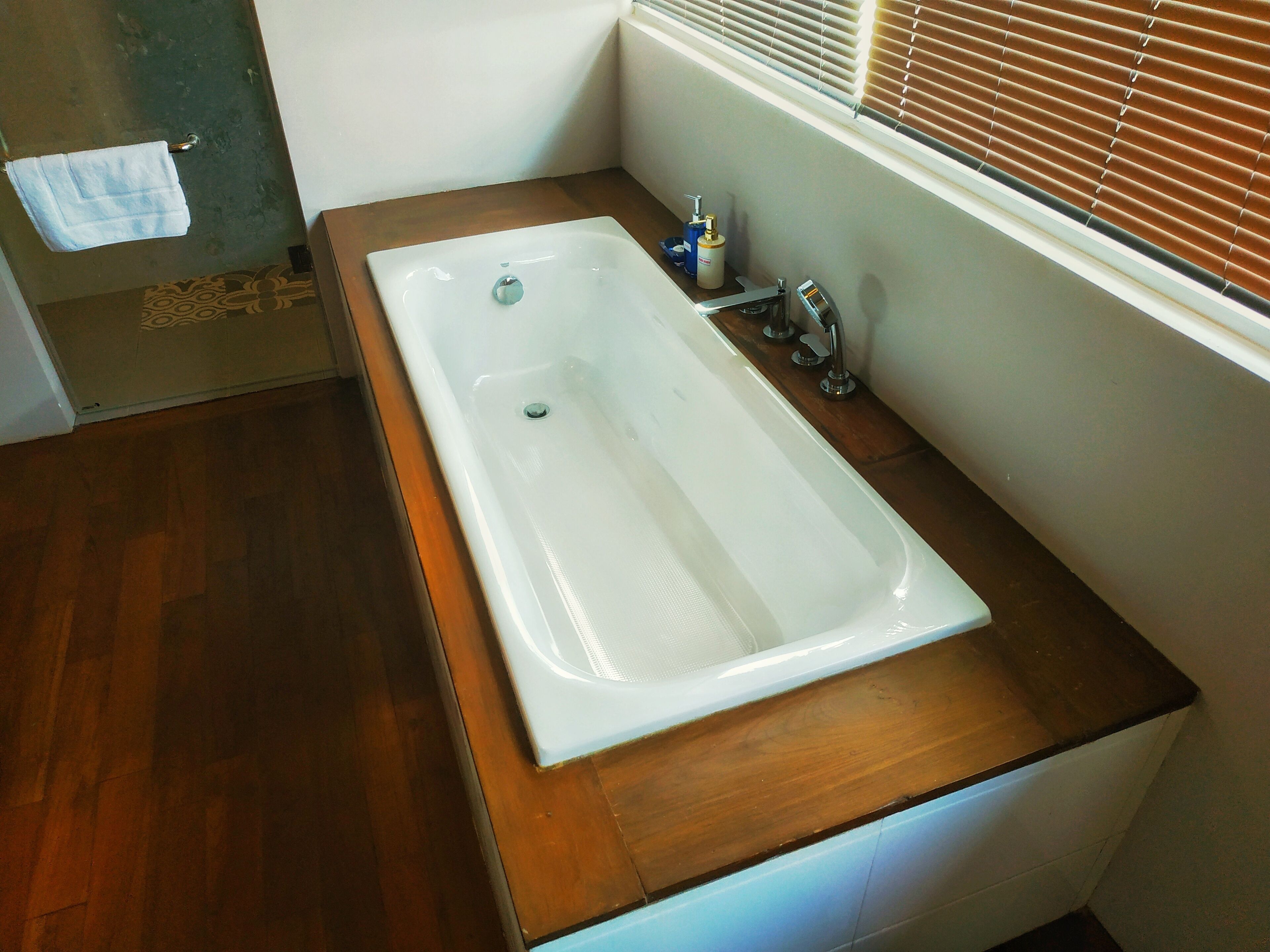 A bathroom with a wooden vanity, a white bathtub, and a window with blinds.