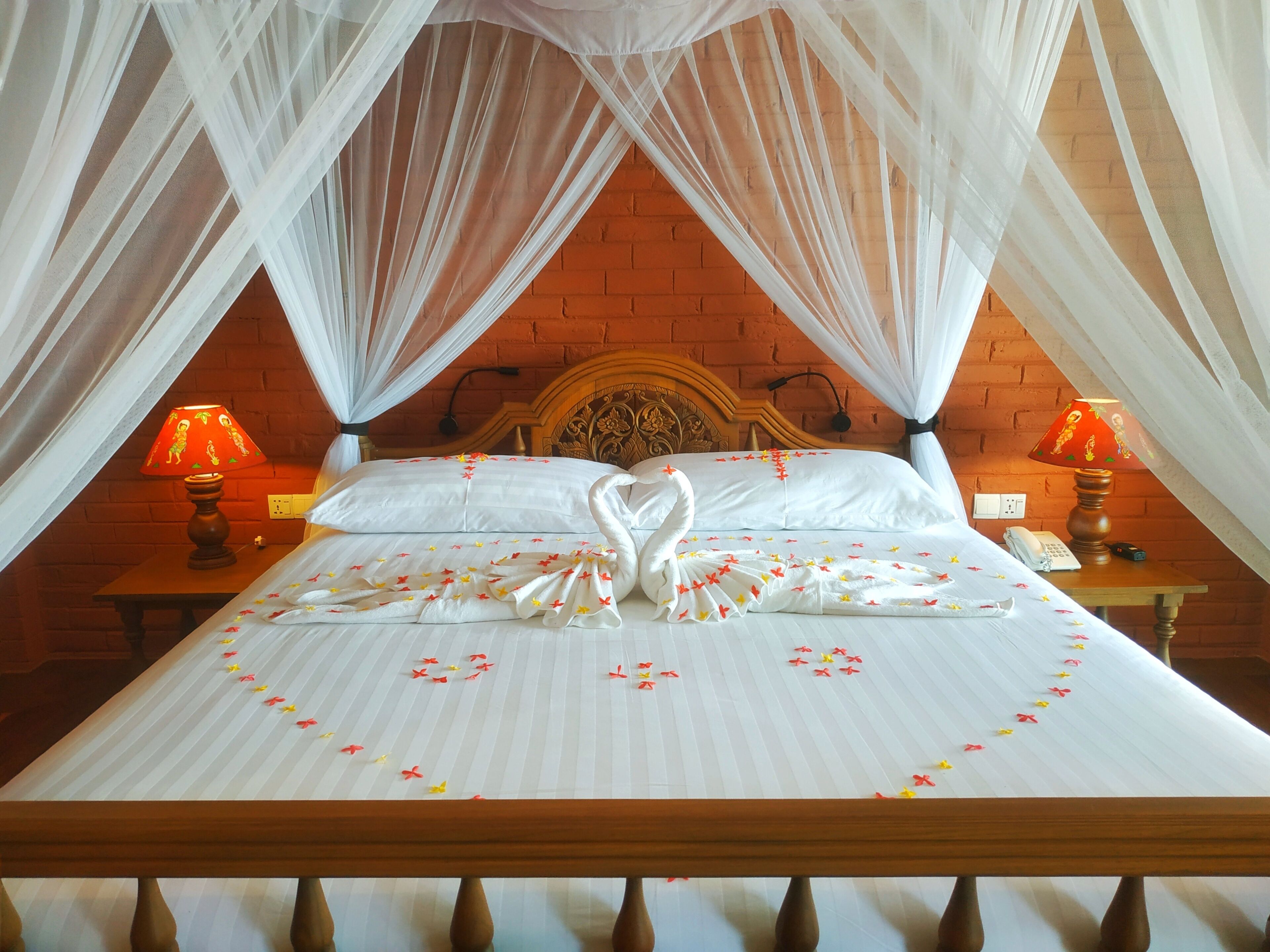 A neatly made bed with a swan-shaped towel, a red and yellow patterned lamp, and a wooden headboard against a brick wall.