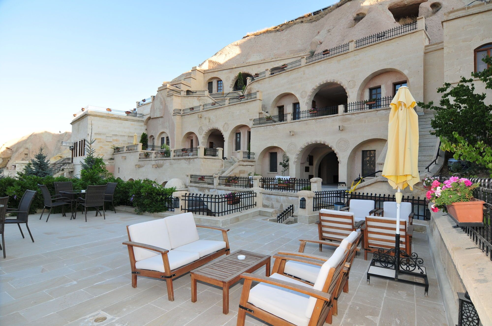 An outdoor seating area with wooden furniture and a yellow umbrella, set against a building with multiple balconies and arches.