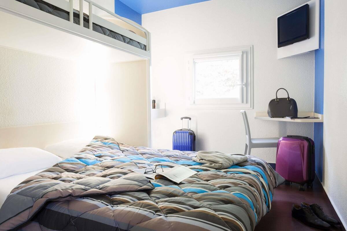 Ein Hotelzimmer mit Etagenbett, einem Koffer, einem pinken Koffer, einem Schuhpaar, einem an der Wand montierten Fernseher und einem Fenster mit Blick auf Bäume.
