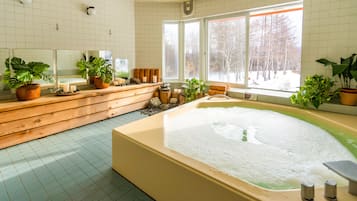 A spa room with a large bathtub, wooden benches, potted plants, and a view of a snowy forest through large windows.