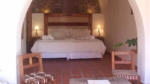 A bedroom with a stone wall, wooden headboard, two bedside lamps, a bed with white linens, and a small table with a chair. - Hostería del Amauta (Purmamarca)