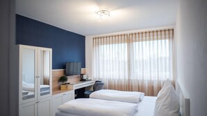 Une chambre d’hôtel moderne, équipée d’un lit, d’un bureau avec une télévision, d’une armoire et d’une fenêtre avec des rideaux légers.