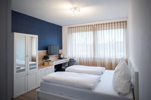 Une chambre d’hôtel moderne, équipée d’un lit, d’un bureau avec une télévision, d’une armoire et d’une fenêtre avec des rideaux légers.