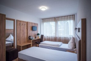 A hotel room with two beds, a wooden wardrobe, a desk with a television, and a window with sheer curtains.