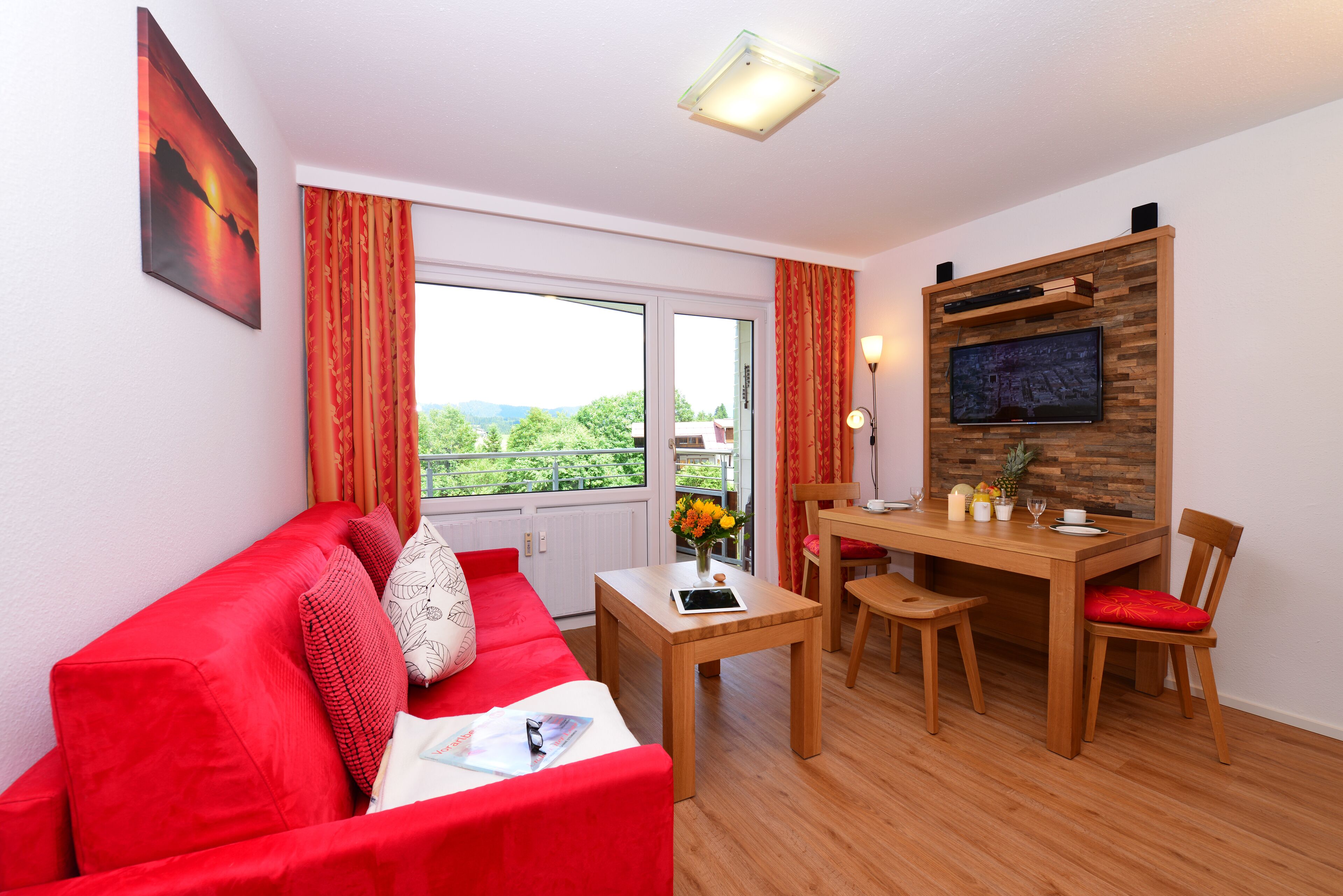 A room with a red sofa, wooden dining table, and a TV mounted on a wooden wall.
