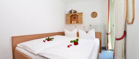 Una cama bien hecha con sábanas blancas y pétalos de rosa roja, un cabecero de madera, un pequeño armario con libros, un reloj de pared y una ventana con cortinas.
