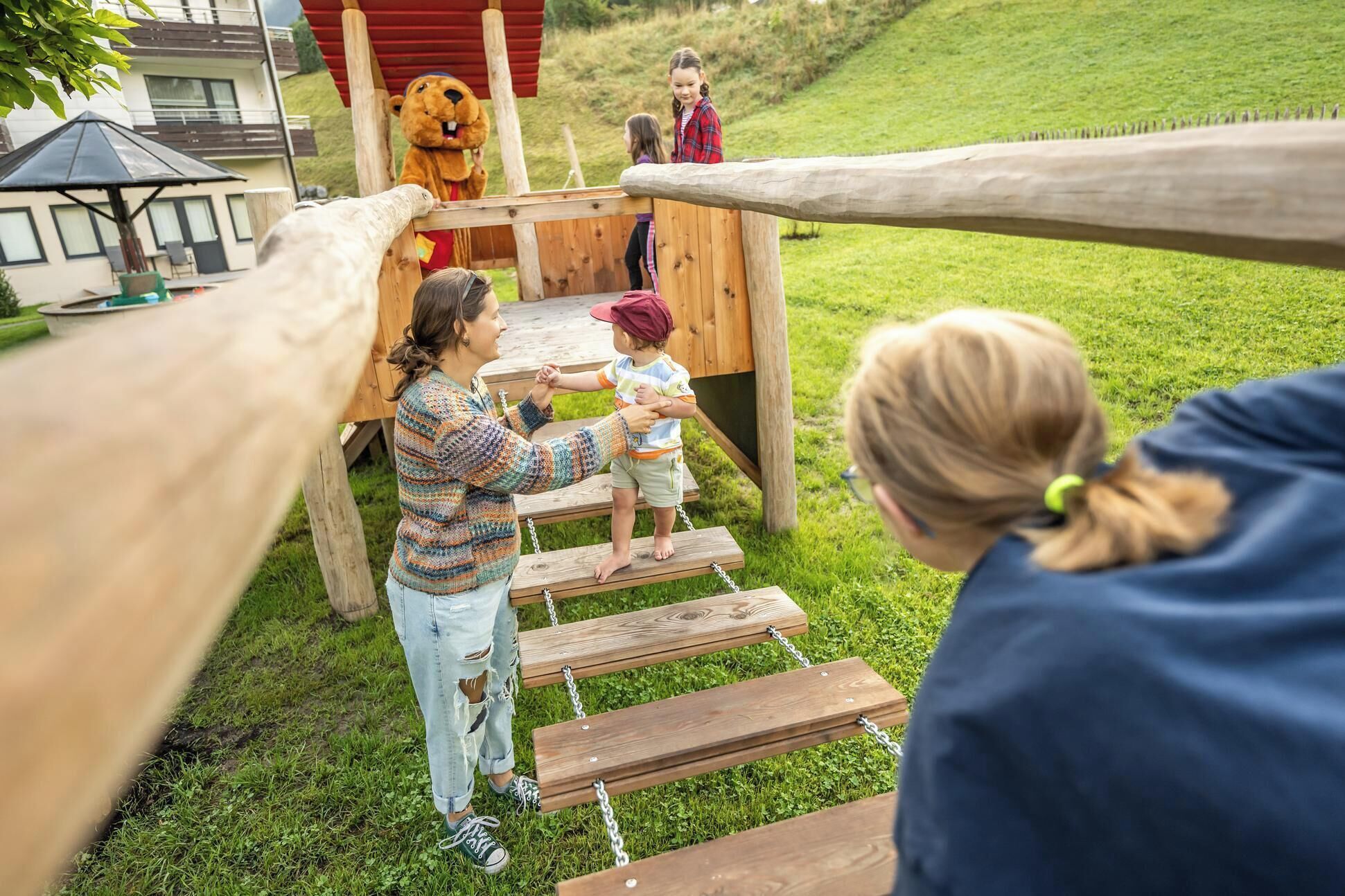 Een houten speeltoestel met een glijbaan en een klimgedeelte, omringd door gras en een gebouw op de achtergrond.