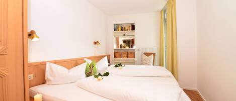 A hotel room with a wooden bed, white bedding, a nightstand with a lamp, a shelf with books and a vase, and a window with curtains.
