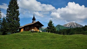 A small wooden chapel with a bell tower, surrounded by a grassy hill and tall trees, with a mountain in the background. - Familienhotel Kleinwalsertal (Mittelberg)