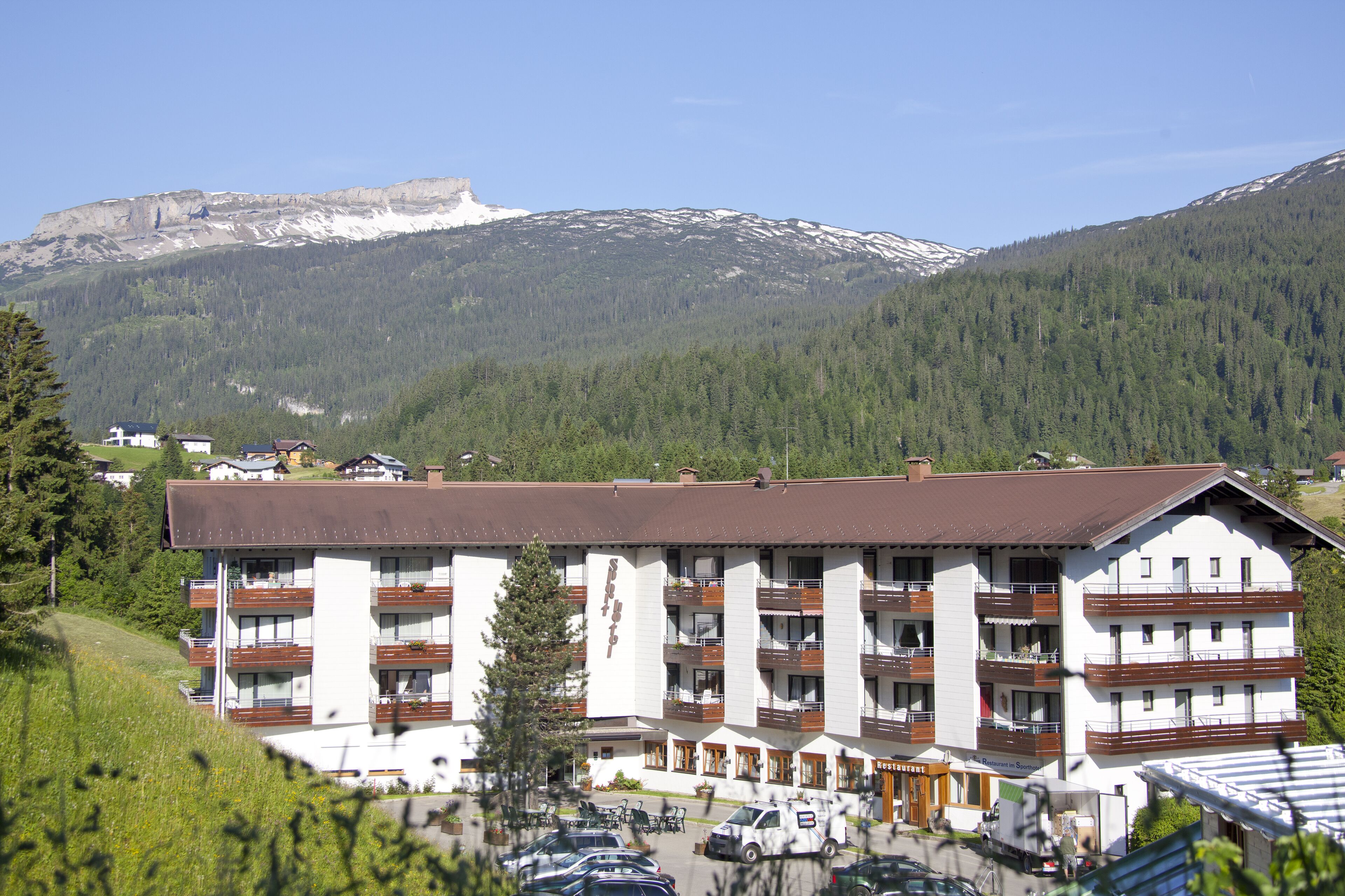 Un hotel de varios pisos con balcones, una zona de estacionamiento y montañas al fondo.
