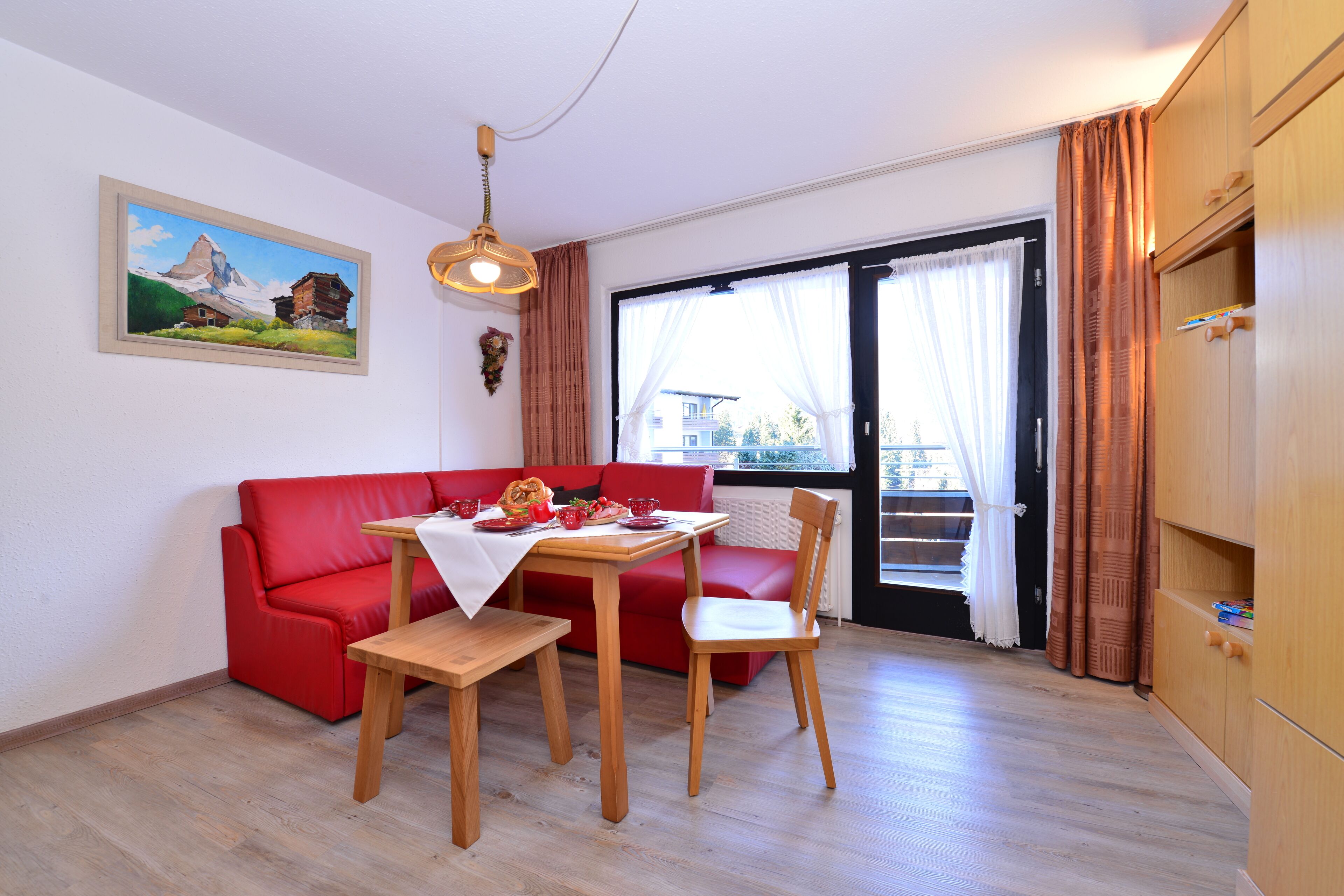 A dining area with a red sofa, wooden table, and chairs. There is a painting on the wall and a window with curtains.