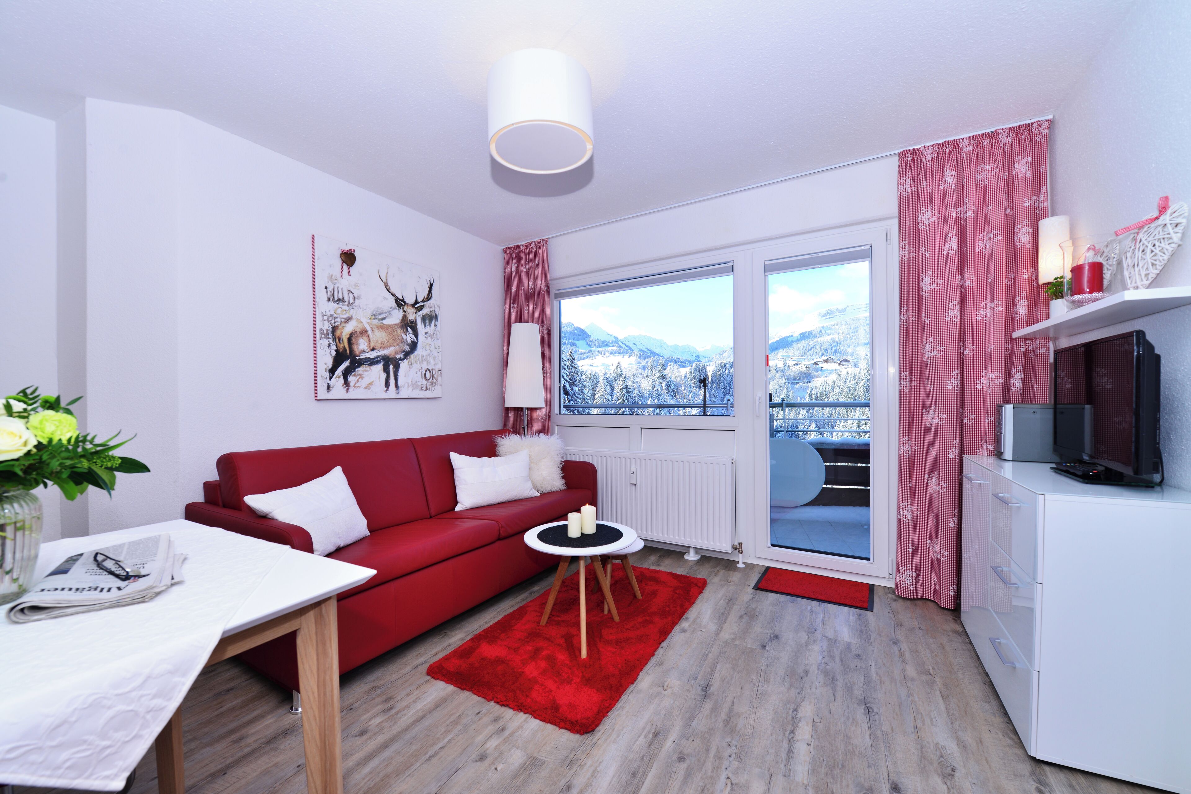 Un salon moderne avec un canapé rouge, une petite table ronde et une vue sur des montagnes enneigées, visible à travers une grande fenêtre.