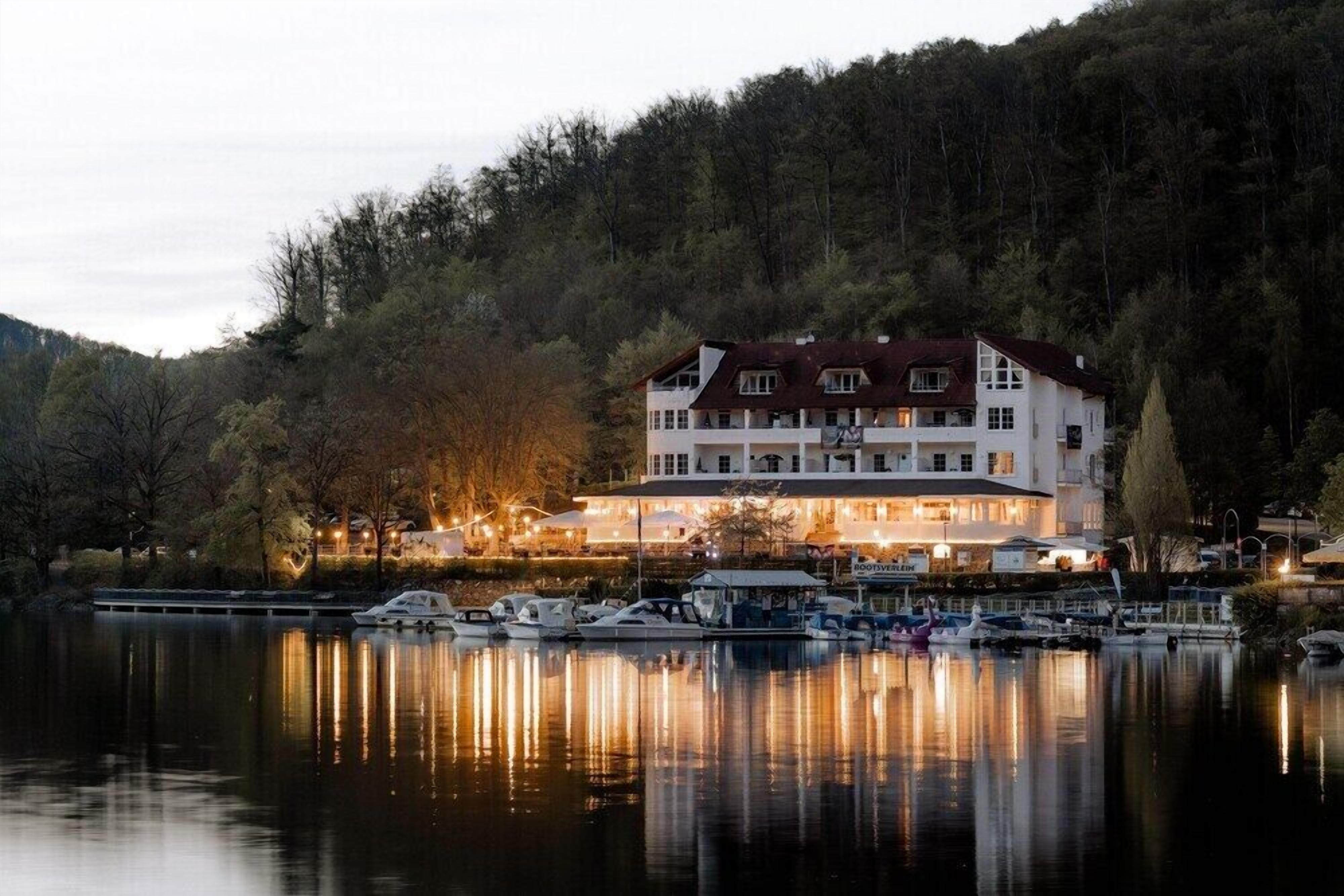 A large white building with multiple stories, likely a hotel, situated by a calm lake with boats docked in front.