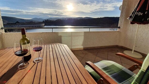 A wooden table with two wine glasses and a bottle, a chair, and a view of a lake and mountains. - Am Edersee (Edertal)