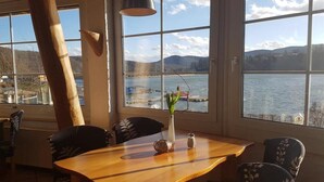 A dining area with a wooden table, chairs, and a large window offering a view of a lake and mountains. - Am Edersee (Edertal)