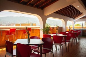 Un restaurante con sillas y mesas rojas, ventanas amplias y vistas a las montañas.